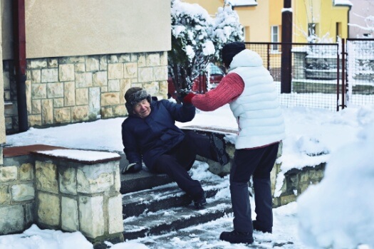 A person helps an elderly man up after he slips on icy, snowy steps.