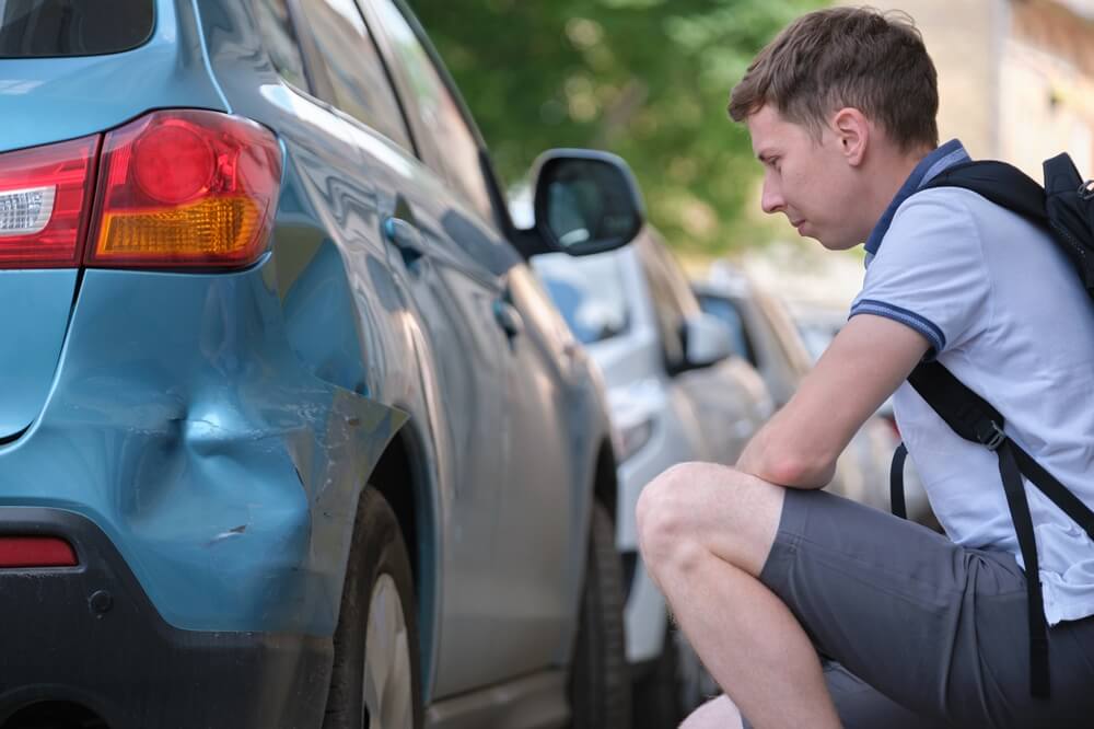 A man sits on the curb examining damage to his car from a hit and run driver - cheap car insurance in Georgia.